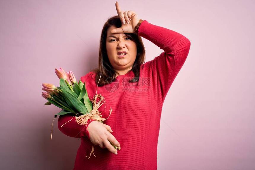 美丽的加上大小的女人拿着浪漫的天然郁金香花束在粉红色的背景上取笑用手指在额头上做失败者手势嘲图片