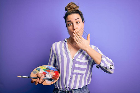 年轻漂亮的金发艺术家女用画笔和调色板在紫色背景上作画图片