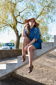 穿着牛仔裙和一顶宽边米色帽子的风趣女孩坐在未完工桥的混凝土台阶上图片