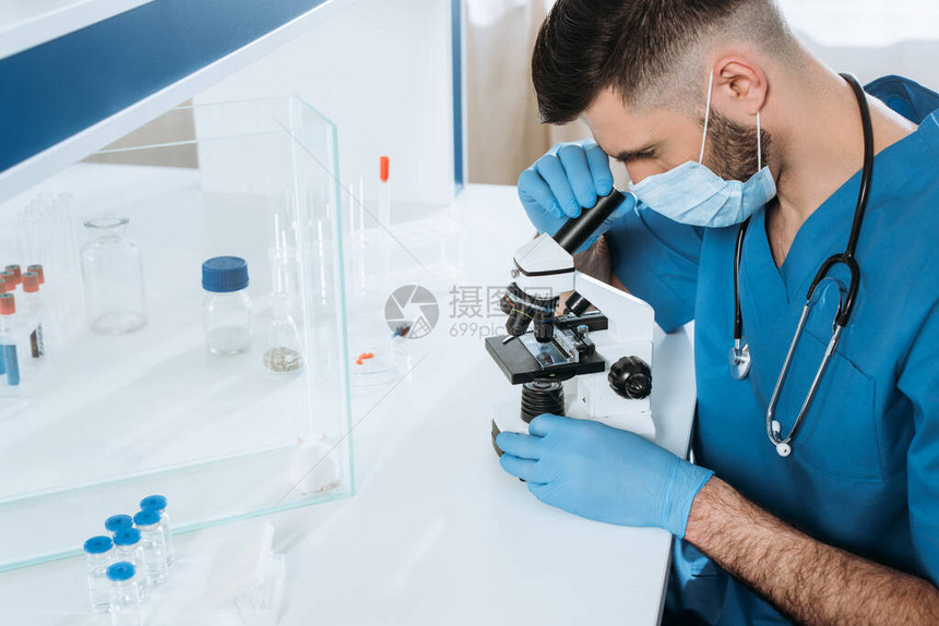 戴着医用口罩和最新手套的年轻生物学家用显微镜在玻璃盒中的白鼠附近和装有药物的容图片