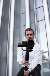 年轻的专业摄像师在3轴云台稳定器上手持专业相机专业设备有助于制作高质量视频而不会晃动摄影师穿着白色连背景图片