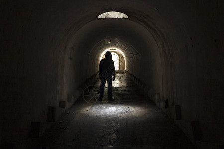 一个身穿深色衣服头戴兜帽的男人正从一条黑暗阴的地下隧道中走到灯的出口图片
