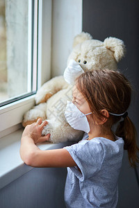 小女孩带着戴面罩抱着泰迪熊在窗户附近图片