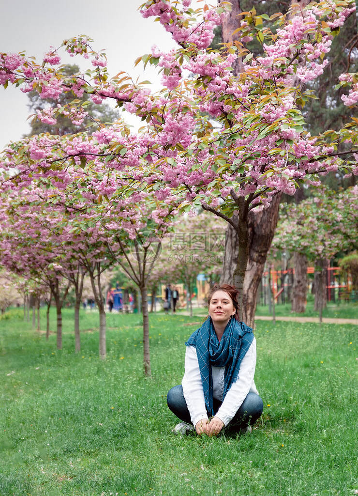 带着牛仔裤白毛衣和蓝围巾的快乐笑容的年轻女子坐在乌克兰基辅市公园中一棵盛开的粉红樱图片