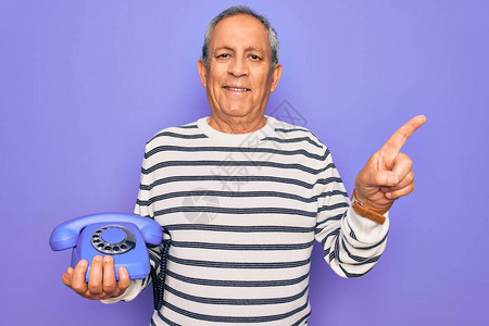 高级英俊的灰色长发男子拿着古老的听筒电话在紫色背景上非常快乐用手和图片