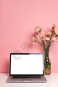 笔记本电脑在白桌上用花瓶中美丽的粉红康乃馨花装饰帕斯特尔玫瑰背景图片