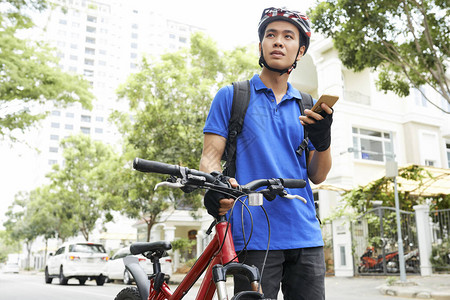 带着智能手机在自行车旁边行走并寻找建筑的越南青年男图片