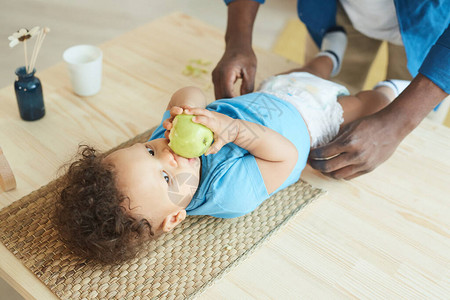 无法辨认的父亲将尿布换成可爱的非裔美国人小婴儿吃苹果和复制空间的高图片