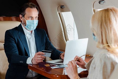 戴医用口罩持信用卡在飞机上靠近女商人时使用笔记本电脑的商人图片