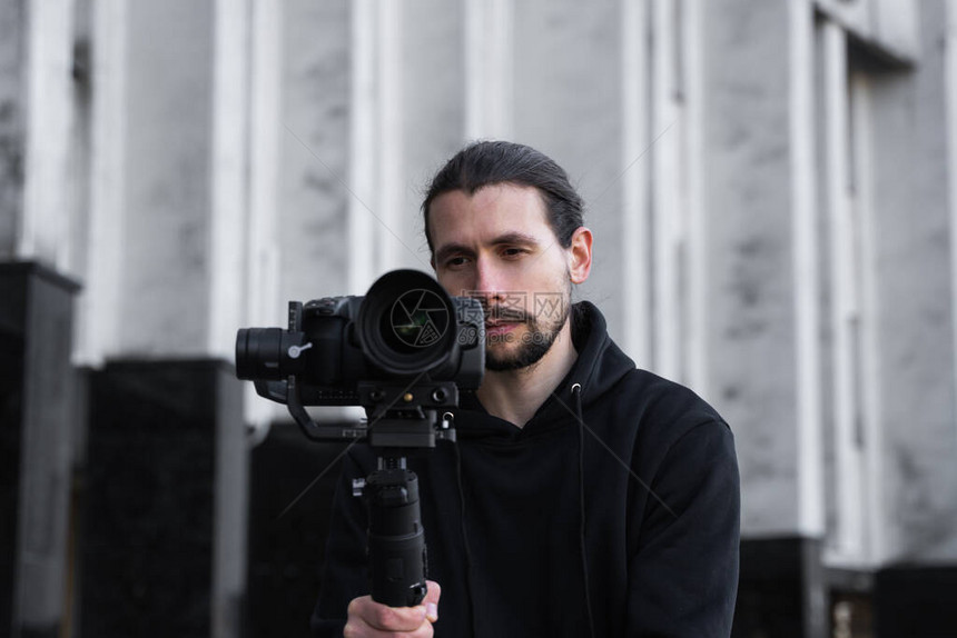 年轻的专业摄像师在3轴云台稳定器上手持专业相机专业设备有助于制作高质量视频而不会晃动身穿黑色连帽衫的摄图片