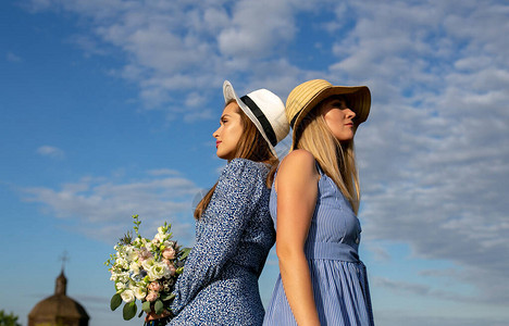 两个身穿蓝色夏裙和帽子的年轻女孩背对着一个美丽的夏日天空和旧教堂站图片