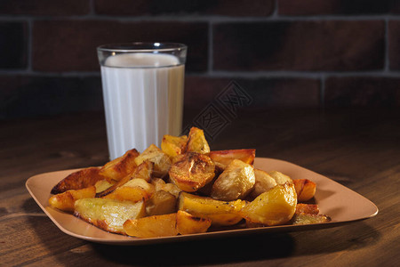 棕色盘子里烤焦的土豆和木制桌子上的切碎玻璃中的牛奶图片
