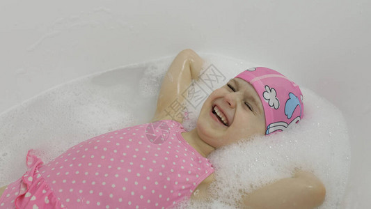 迷人的四岁女孩穿着粉红色泳装和泳帽用沐浴泡沫洗澡躺在浴缸里可爱的金发孩子的卫生可爱的女孩微笑浴室里背景图片