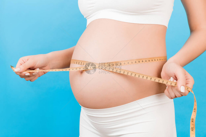 孕妇穿着白色内衣的裁剪图像测量她的腹部以检查婴儿的发育在蓝色背景的厘米卷尺健康怀图片