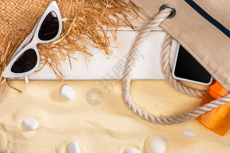 智能手机和防晒霜的顶部视图靠近草帽的袋子和白色木板上的太阳镜和沙图片