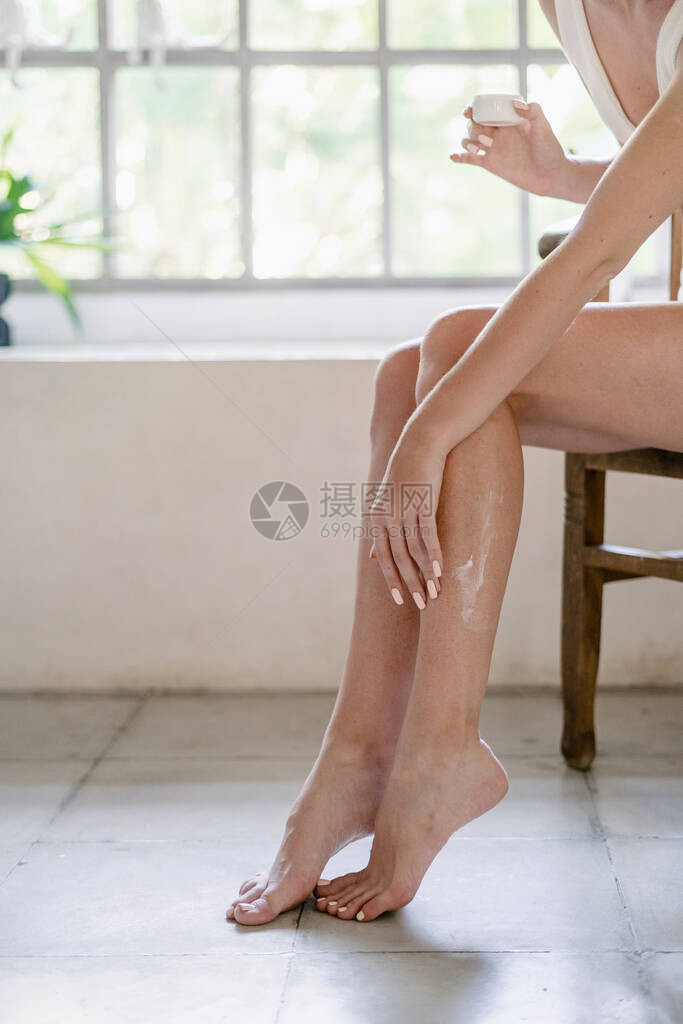 天然护肤理念年轻成年女孩在腿上涂抹乳液霜坐在白色浴室的椅子上手持装有保湿产品的容器的裁图片