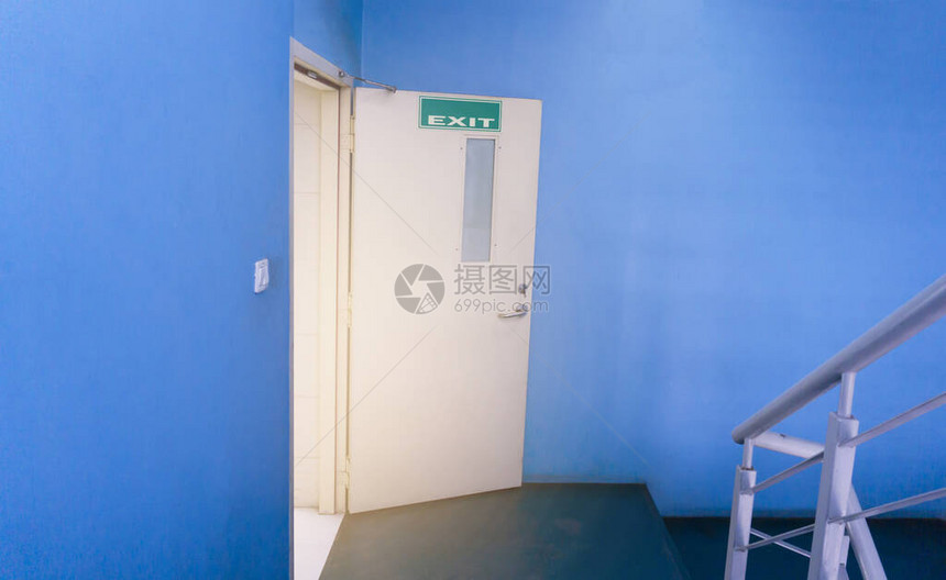 一扇奶油色的消防出口门从一间蓝色的墙壁房间打开到一个带有灰色栏杆的楼梯图片
