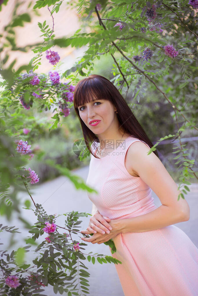 穿着粉红无袖的夏裙的深黑头发年轻女子在绿和鲜花植物间的一图片