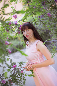 穿着粉红无袖的夏裙的深黑头发年轻女子在绿和鲜花植物间的一图片
