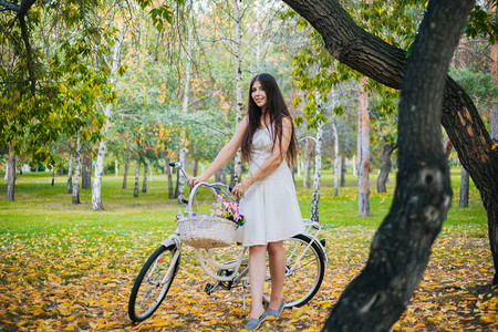穿着白裙子的女孩和穿自行车的背心以及一篮子鲜花在晚上秋天公园黄图片