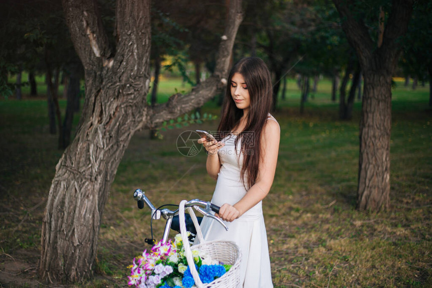 身穿白裙子的女孩和手拿着智能手机的背心站在一辆自行车旁边图片
