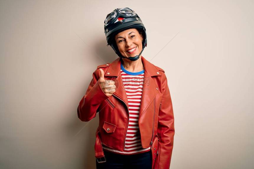 中年摩托车手妇女戴着摩托车头盔和夹克在白色背景做快乐的拇指手势用手看着相机显示成图片