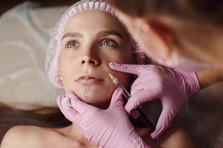 在美容诊所填充剂注射中进行年轻化程序的妇女她平静地躺在诊所里戴着粉色手套的专业美容师正在用透明质酸填充女皱背景图片