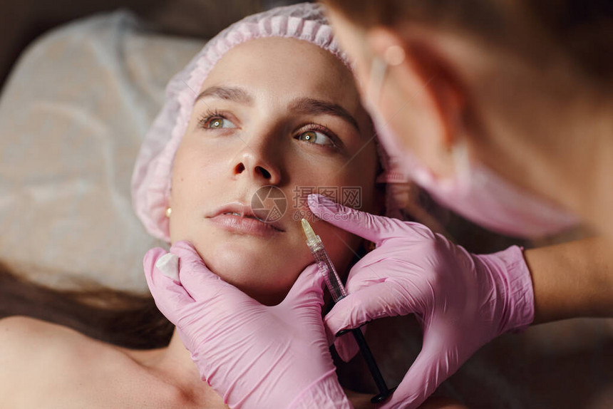 在美容诊所填充剂注射中进行年轻化程序的妇女她平静地躺在诊所里戴着粉色手套的专业美容师正在用透明质酸填充女皱图片