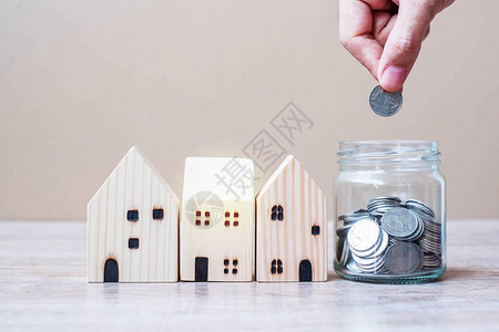 男人手把硬币放在玻璃罐子里和桌子背景上的木屋模型商业投资省钱房地产和图片