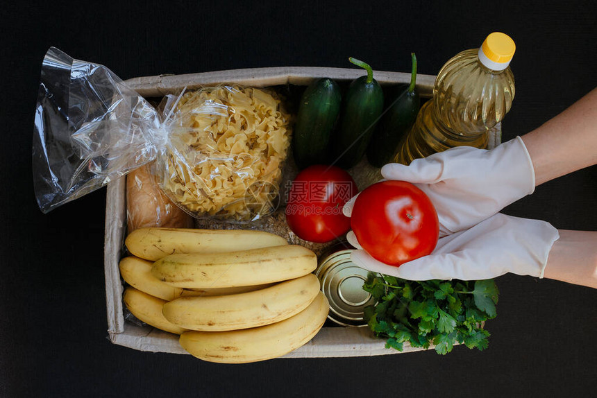 一只戴着橡胶手套的手拿着番茄一个装有食物的捐款箱水果蔬菜罐头食品面食和葵花籽油装在盒子里食物方图片