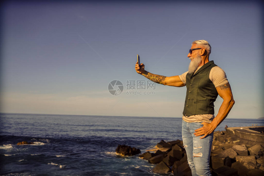 时髦的老人在海边使用智能手机应用程序成熟的时尚男享受新趋势技术的乐趣科技和快乐的老年生活方式概念专注于图片