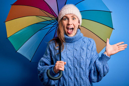 年轻漂亮的金发女郎在孤立的蓝色背景上举着五颜六色的雨伞图片