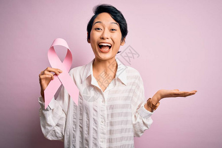 年轻漂亮的亚洲女孩在孤立的背景上举着粉色癌症丝带符号图片
