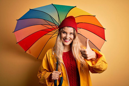 身穿雨衣和羊毛帽的金发年轻美女拿着多彩大雨伞笑得开心图片