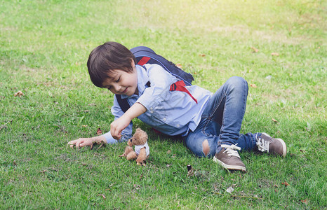 可爱的小男孩背着包躺在草地上和泰迪熊在公园玩耍图片
