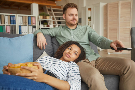 年轻混合种族夫妇在家里看电视吃零食在舒适沙发上放松复图片