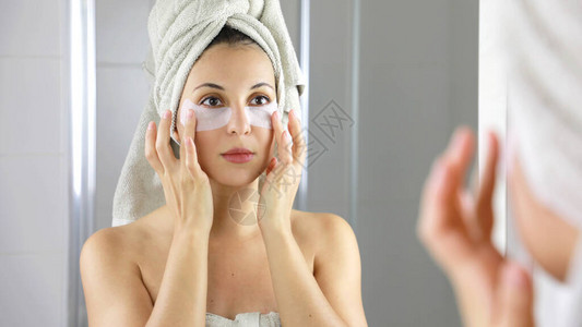 美丽的女人应用抗疲劳眼罩在浴室的镜子里看着自己皮肤护理女孩在眼睛下方触摸织物面膜贴背景图片