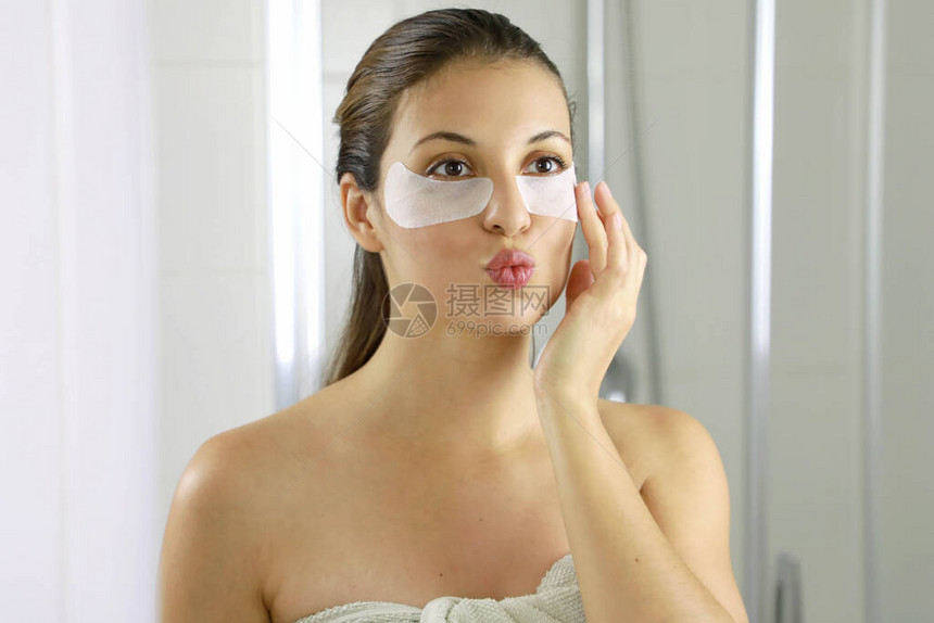 有吸引力的女人应用抗疲劳眼罩在浴室的镜子里看着和亲吻自己皮肤护理女孩在眼睛下方触摸织物面膜贴图片
