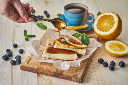 美味的早餐煎饼配蜂蜜蓝莓橙子薄荷和一杯茶一个茶匙的人将蜂蜜倒在煎饼上图片
