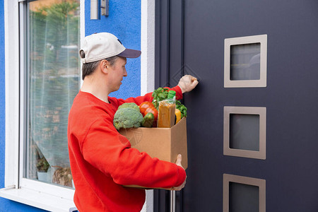 一位身穿红色毛衣和帽子的送餐员或志愿者正拿着一盒产品敲门图片