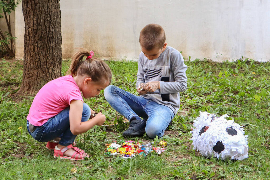 两个孩子在花园的地上吃着像足球一样的破皮纳塔玩具糖果女孩和男孩蹲在草地上拆糖果特写图片