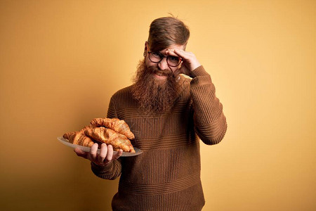 留着胡须的爱尔兰红头发男子在黄色背景下吃法式羊角面包糕点图片