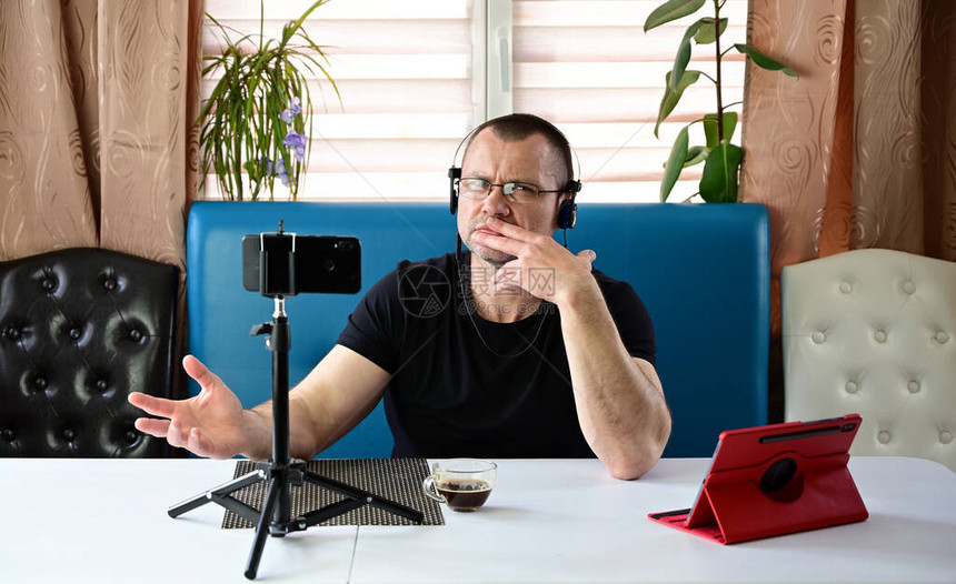 一个戴着耳机和眼镜的成年男子在家里通过智能手机和平板电脑举行视频会议时说话的肖像一位模特穿着黑色T恤坐在图片