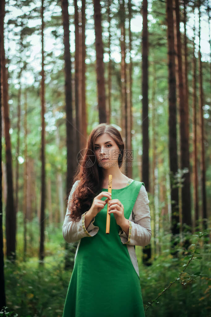 身穿绿色中世纪连衣裙的女人站在松树背景下的树林中吹奏木笛阴暗中的女孩在手工烟斗上演奏音乐阴暗图片
