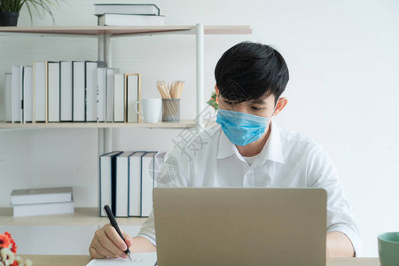 一名年轻的亚洲男子戴着医用口罩在家工作与社会保持距离以防止冠图片