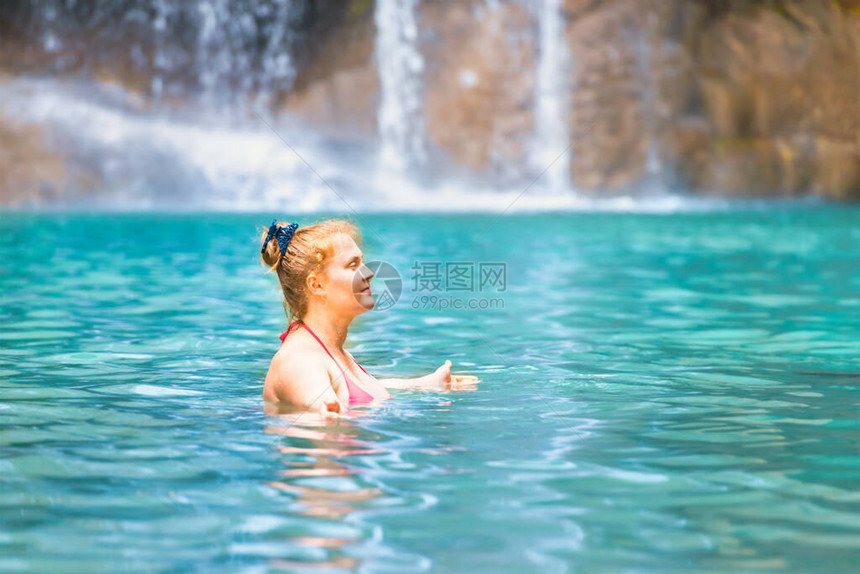 身穿粉色比基尼泳装的红发年轻女子在有瀑布的翡翠热带湖中放松四面佛公园图片