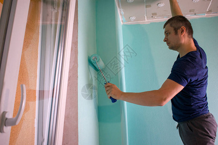 画家用油漆覆盖墙壁师父在公寓里当油漆滚筒男人用水溶涂料在薄荷色的墙上涂漆维修工作房间内墙背景图片