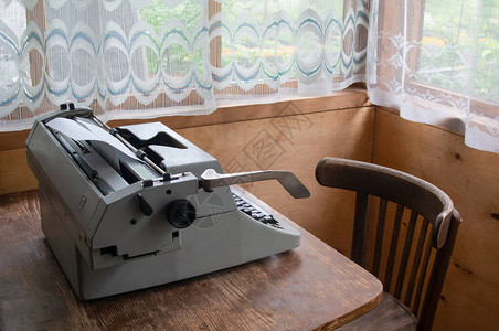 Retro打字机站在一个旧椅子旁边的一栋有大窗户的木房中的图片