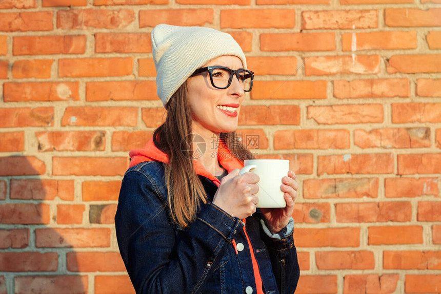 带着白帽子和牛仔夹克的砖墙背景上拿着白色杯子的年轻微笑女青年戴着图片