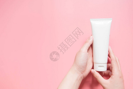 年轻的女手拿着粉红色背景上的空白色挤压瓶塑料管奶油乳液凝胶面部泡沫或护肤品的包装化妆品美容产品牌模背景图片